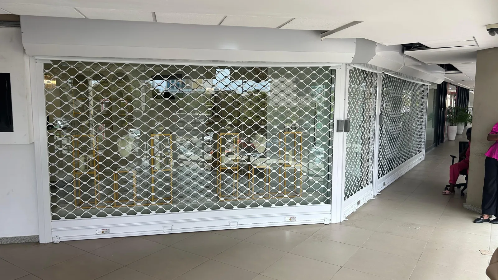 Grade de enrolar tipo lagarto branca, em malha de elos, à frente de uma loja numa galeria comercial — Grupo KEFE, Luanda.