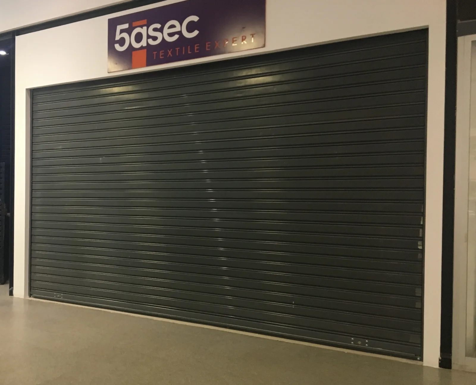 Grade de enrolar cega cinzenta-escura, fechada, numa loja de lavandaria 5àSec num centro comercial — Grupo KEFE, Luanda.