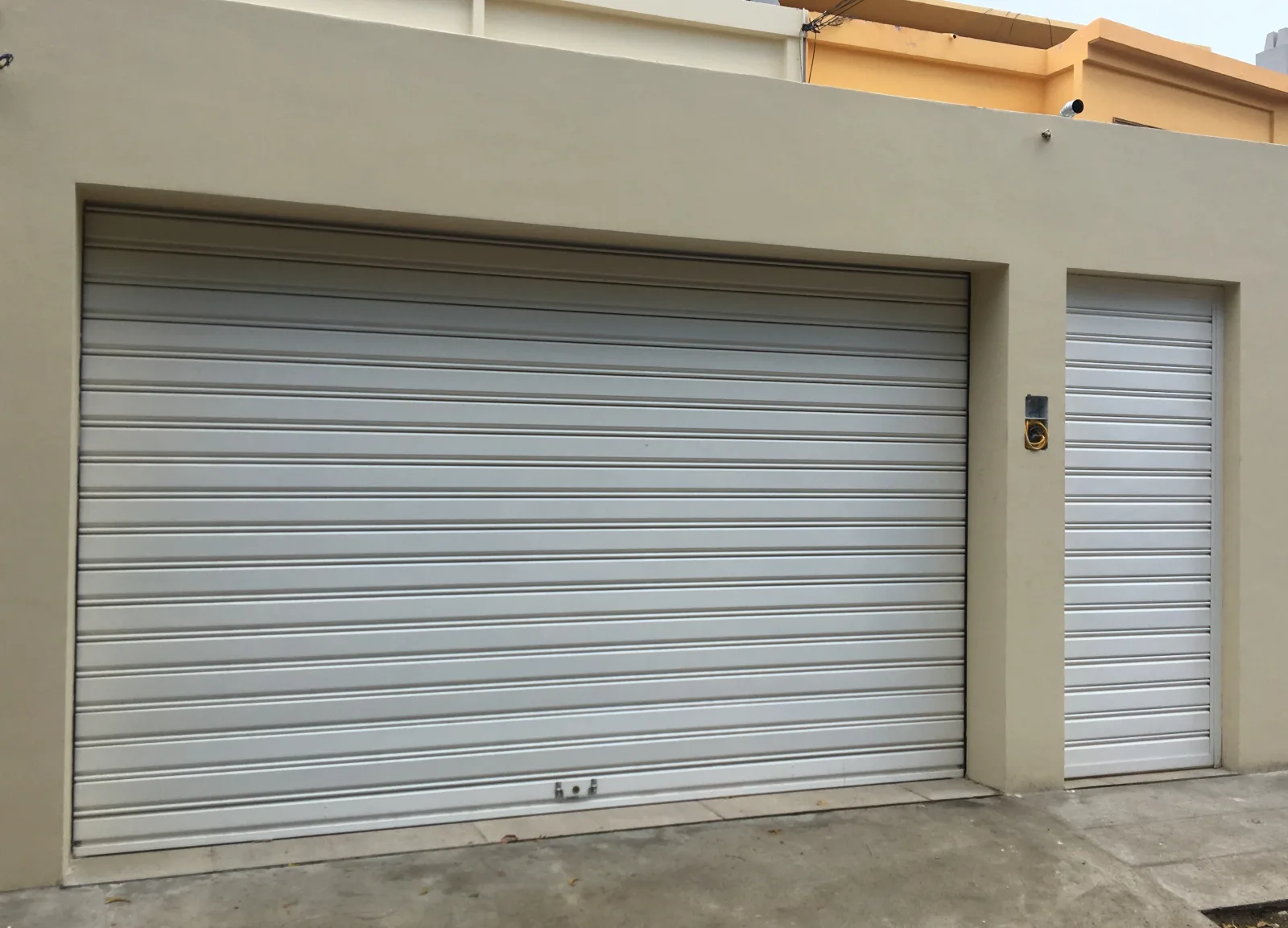 Duas grades de enrolar cegas claras — de garagem e de porta — na fachada de uma moradia — Grupo KEFE, Luanda.