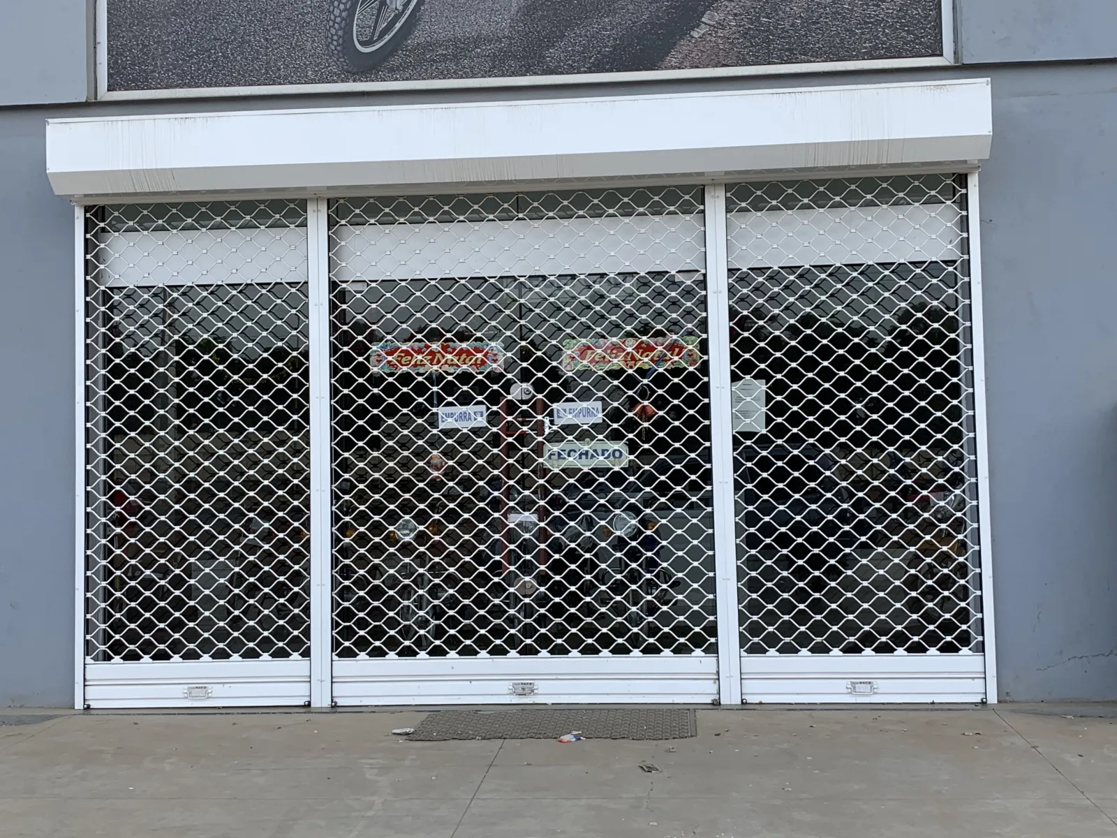 Grade de enrolar transparente, de malha metálica, fechada à entrada de uma loja com montra — Grupo KEFE, Luanda.