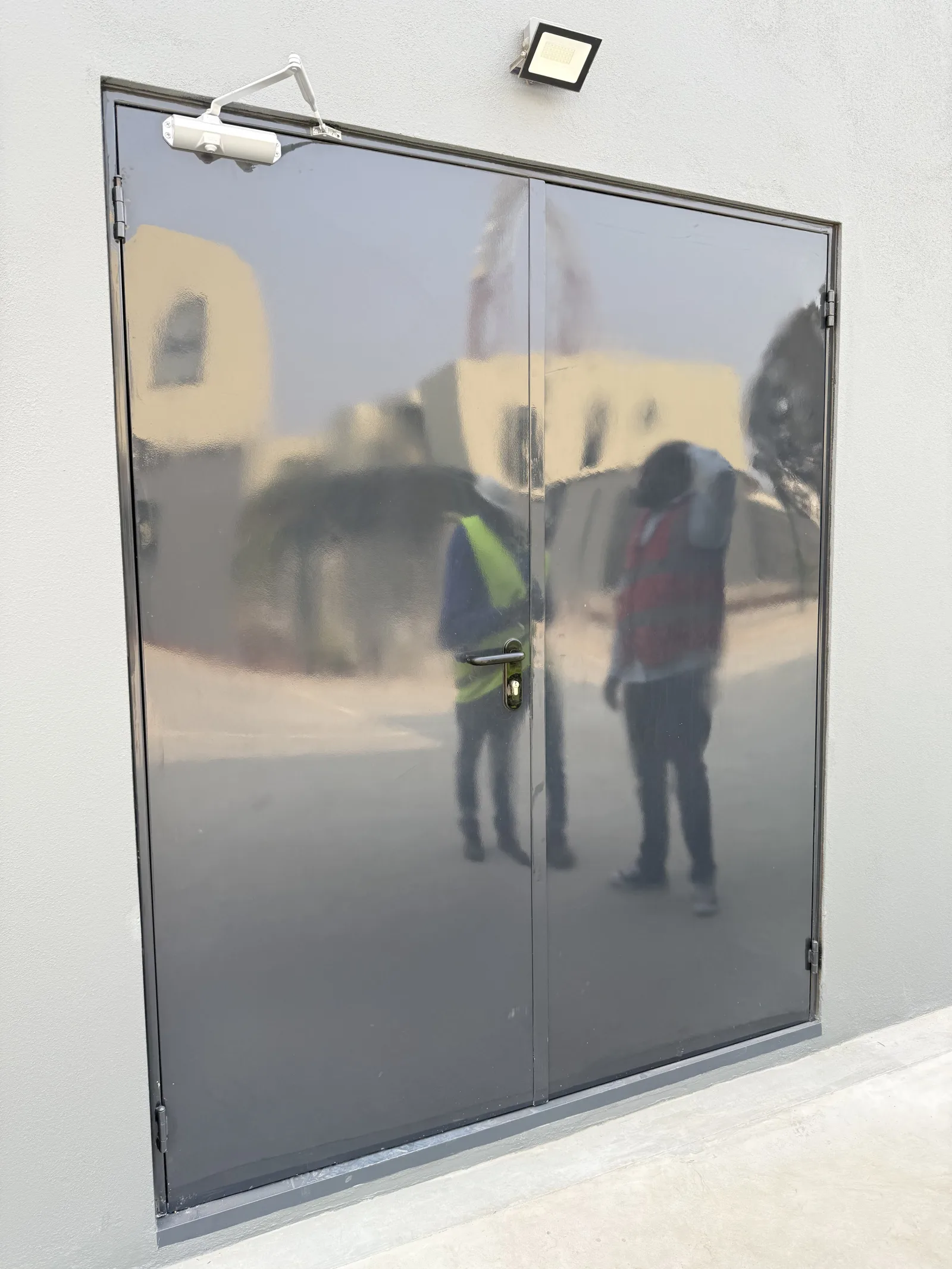 Porta corta-fogo cinzenta de duas folhas com mola de fecho automático, no exterior de um edifício — Grupo KEFE, Luanda.