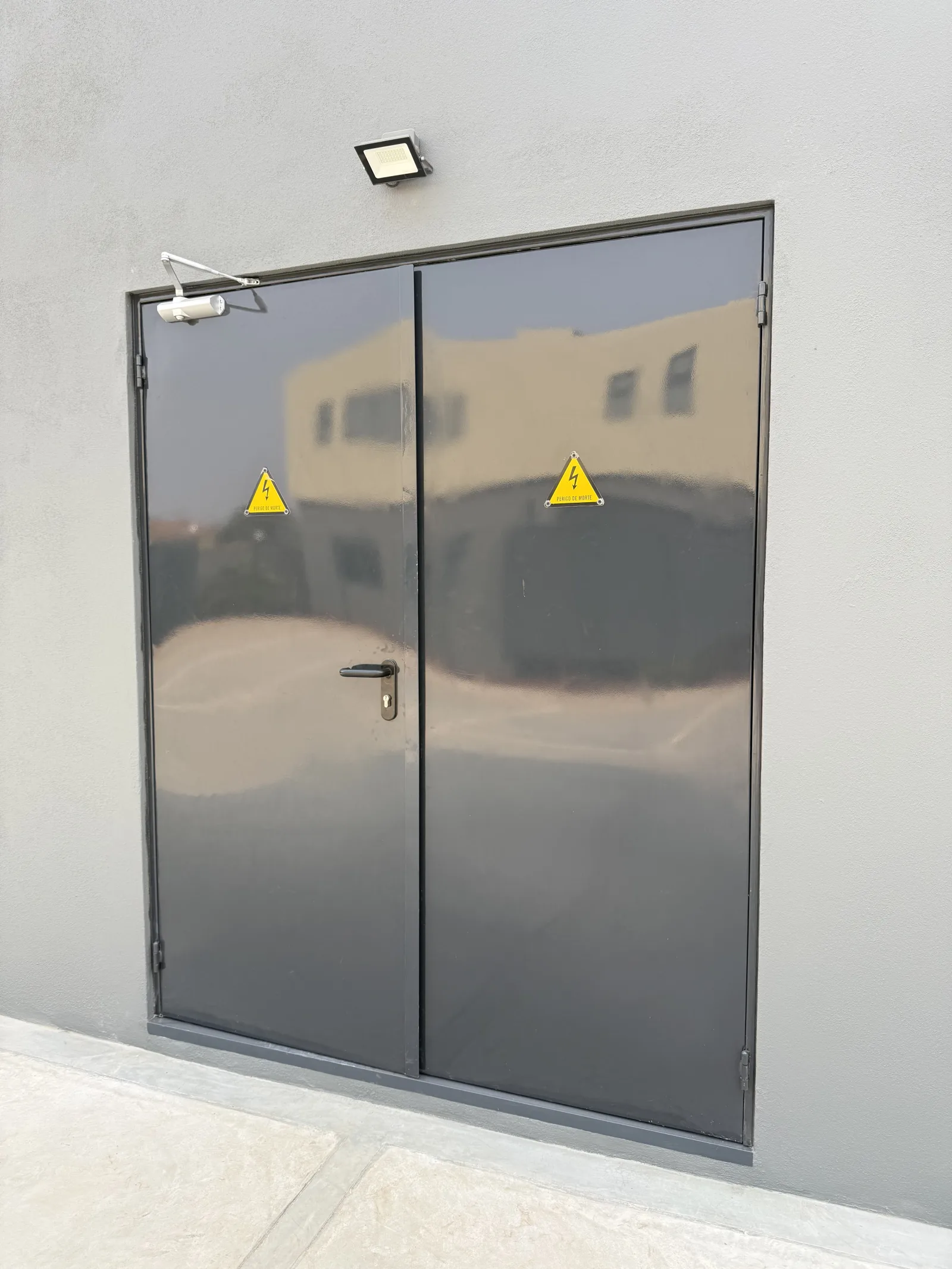 Porta corta-fogo cinzenta de duas folhas com sinalização de perigo elétrico, numa casa de máquinas — Grupo KEFE, Luanda.