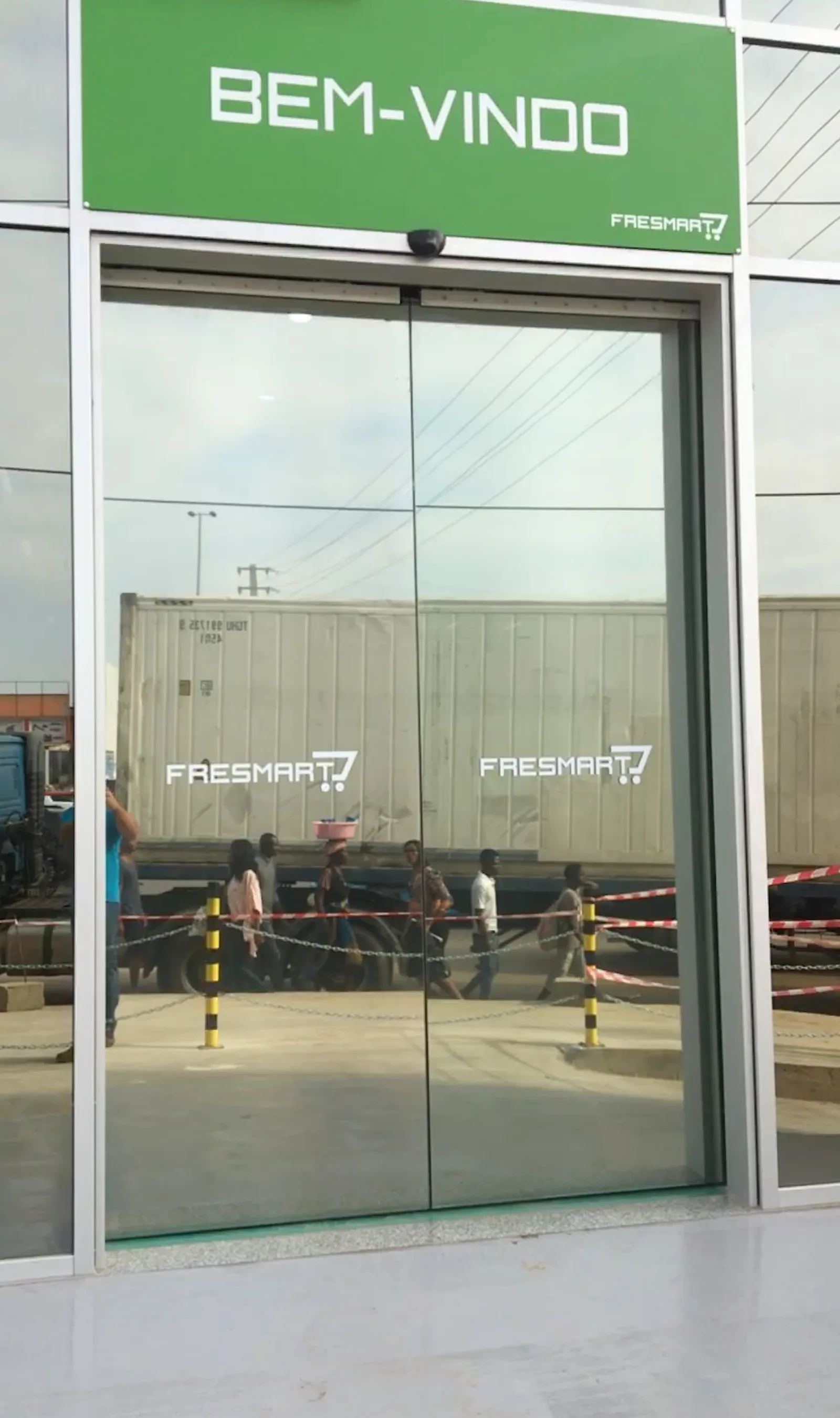 Porta automática de vidro corrediça à entrada do supermercado Fresmart, sob o letreiro "Bem-vindo" — Grupo KEFE, Luanda.