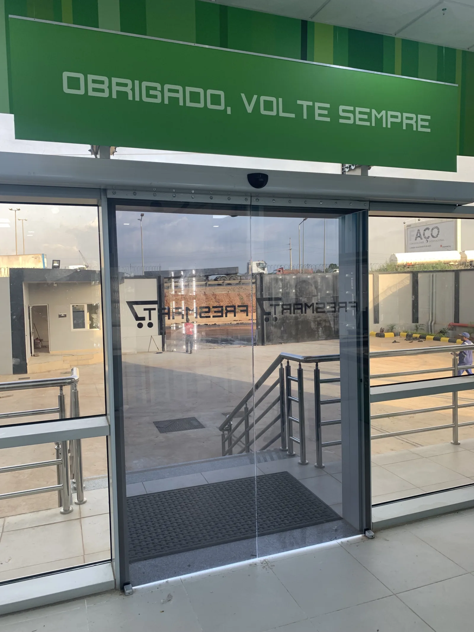 Porta automática de vidro na saída do supermercado Fresmart, sob o letreiro "Obrigado, volte sempre" — Grupo KEFE, Luanda.