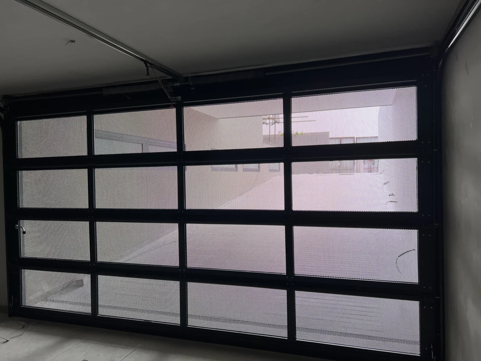 Portão seccionado de estrutura preta e painéis em rede a deixar passar a luz, visto do interior da garagem