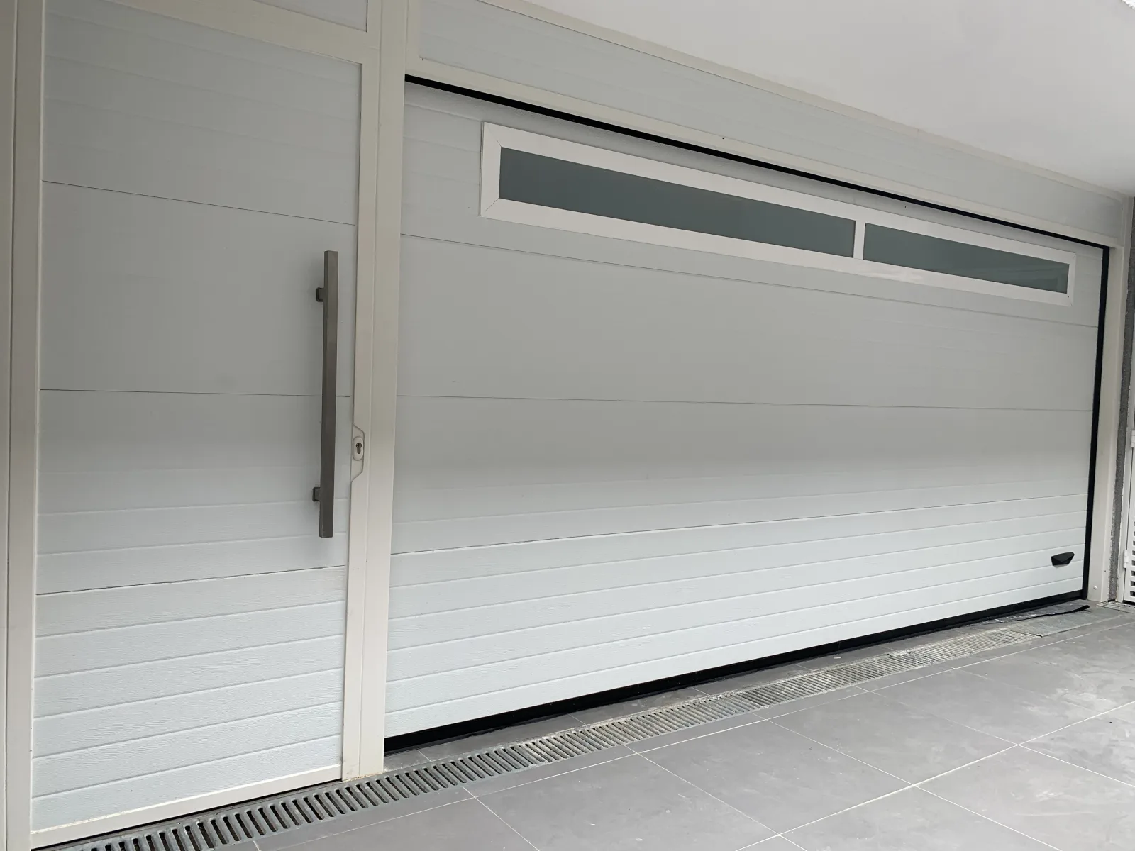 Portão seccionado branco de painéis lisos com faixa de janelas no topo e porta de serviço ao lado