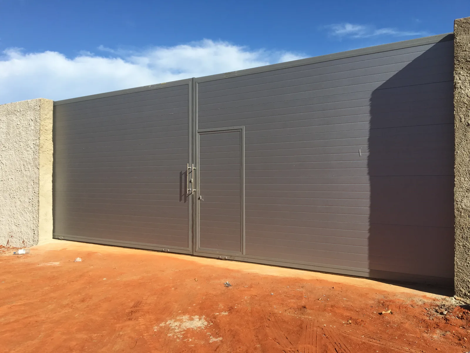 Portão de correr em chapa cinzenta lisa, a fechar o muro de um lote — Grupo KEFE, Luanda.