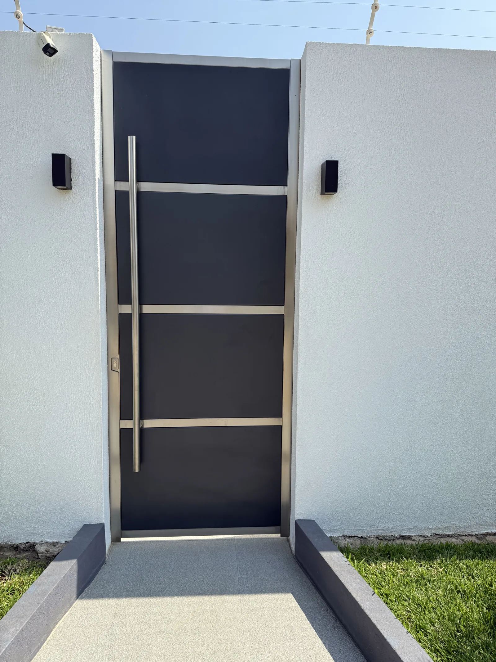 Porta de entrada pivotante, em painel metálico escuro com puxador vertical, numa moradia — Grupo KEFE, Luanda.