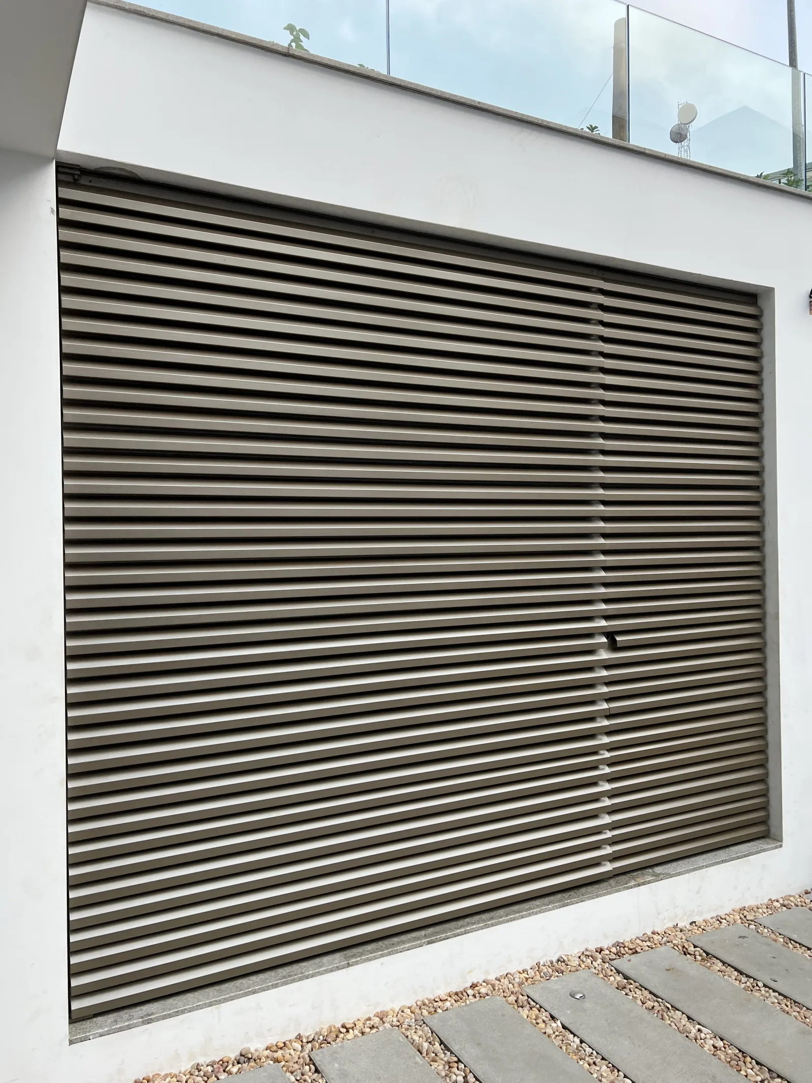 Portão de garagem em lâminas metálicas horizontais, bege, com guarda de vidro no piso superior — moradia, Grupo KEFE, Luanda.