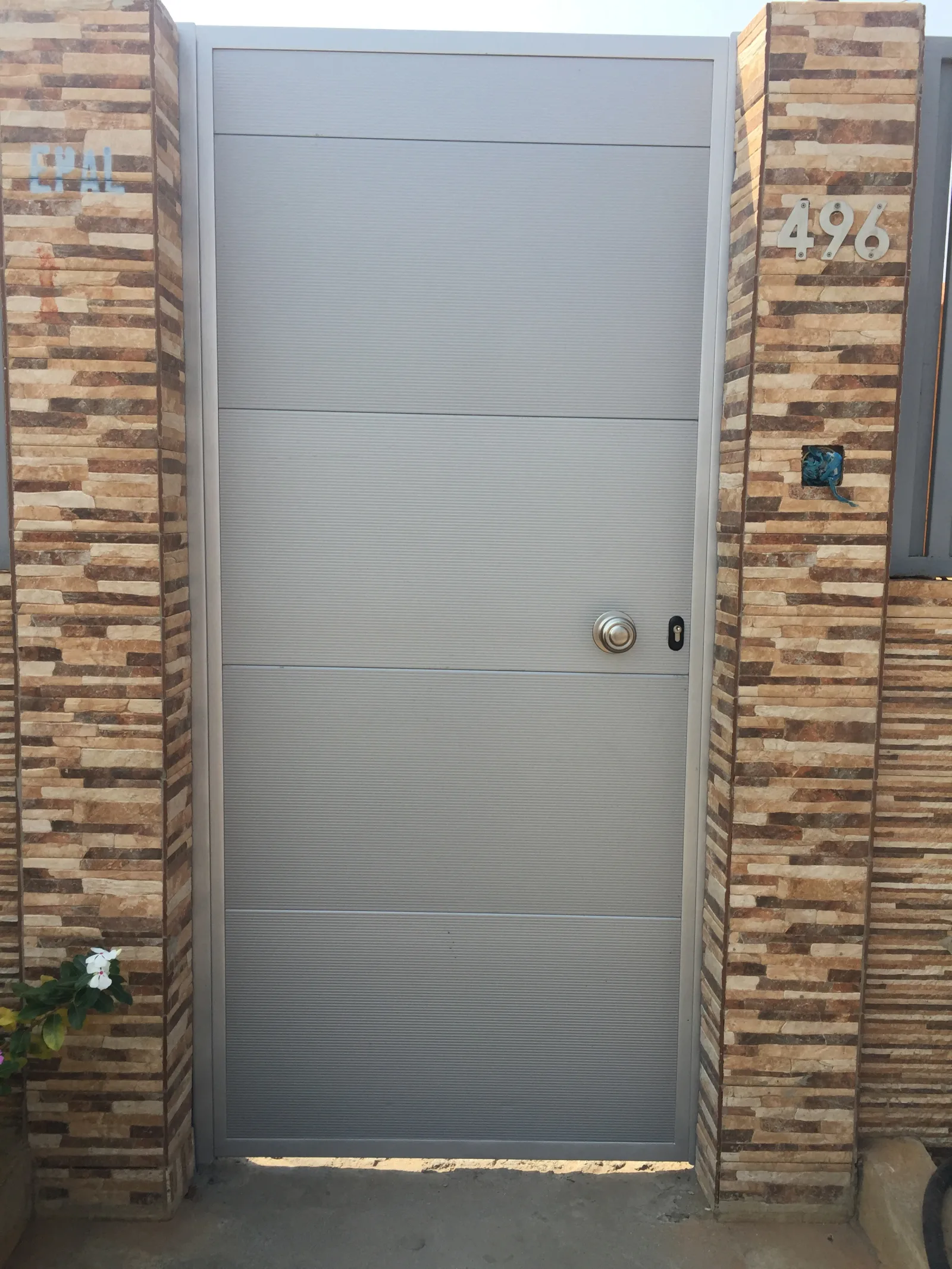 Porta de entrada pedonal cinzenta, com frisos horizontais, num pórtico revestido a tijolo — Grupo KEFE, Luanda.