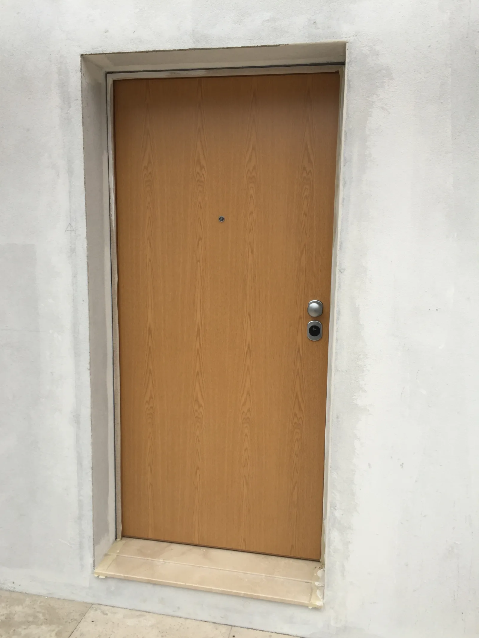 Porta de segurança com acabamento imitação de madeira clara, instalada numa parede rebocada — Grupo KEFE, Luanda.