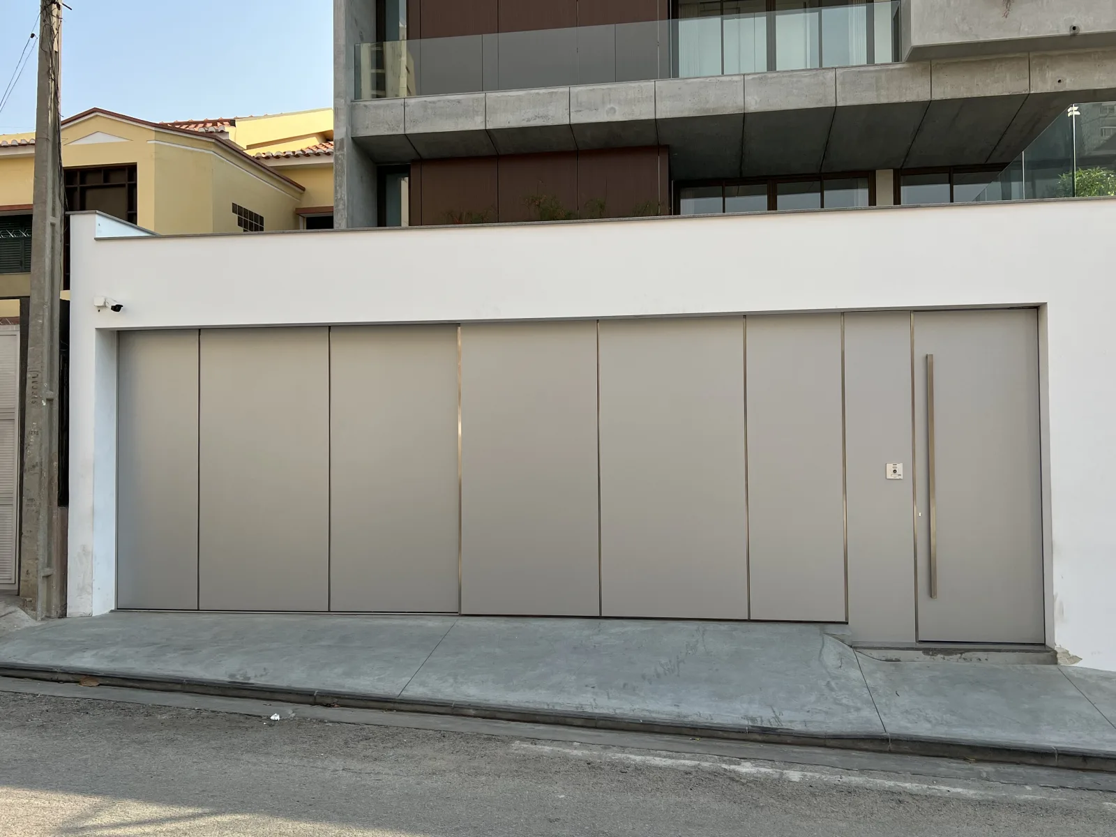 Portão de correr largo, em chapa metálica cinzenta, à entrada de uma moradia moderna — Grupo KEFE, Luanda.