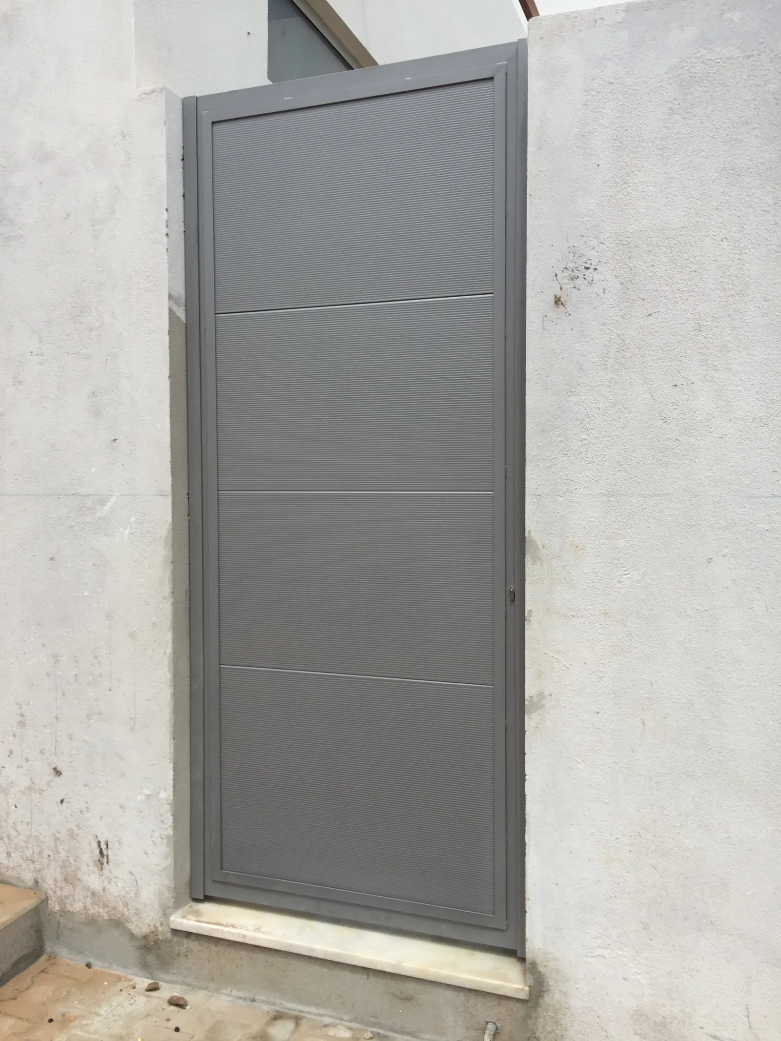 Porta pedonal metálica cinzenta, de painel liso, instalada numa parede rebocada — Grupo KEFE, Luanda.