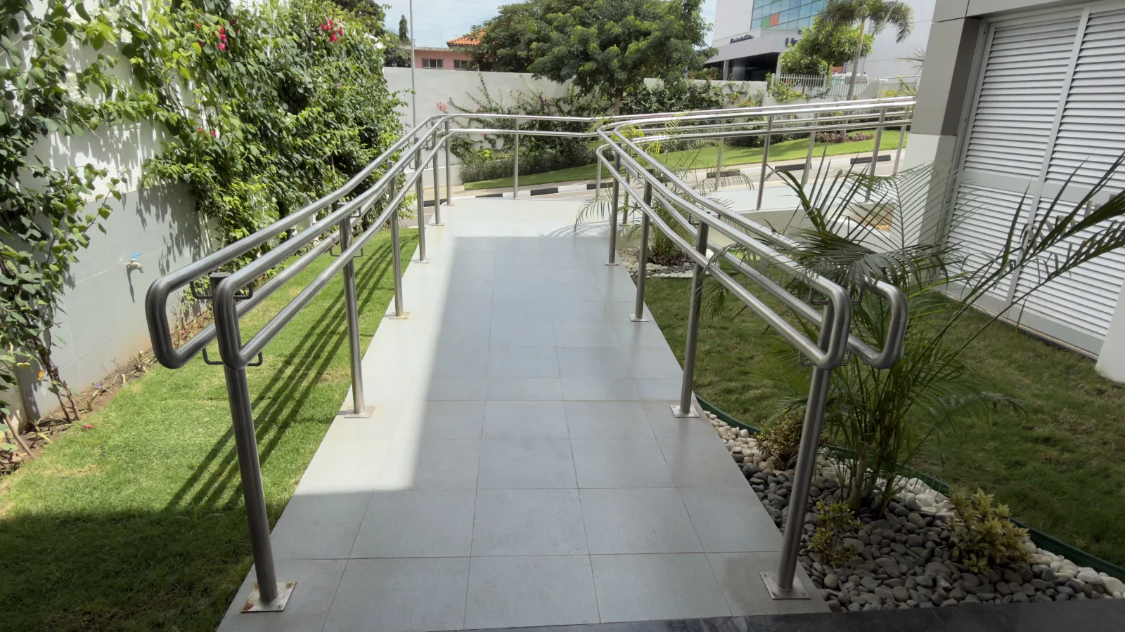 Rampa de acesso com corrimão de aço inoxidável e relvado, junto a um edifício — Grupo KEFE, Luanda.